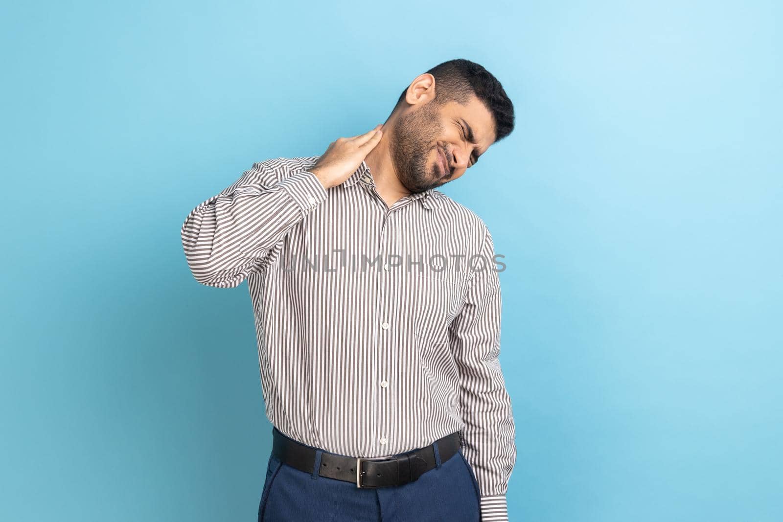 Anxious upset businessman massaging numb neck feeling pain, muscles tension, uncomfortable sleeping conditions, wearing striped shirt. Indoor studio shot isolated on blue background.