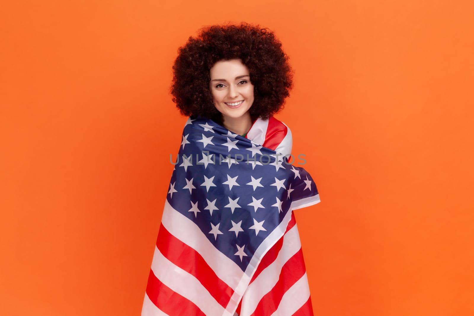 Satisfied woman with Afro hairstyle wearing green casual style sweater standing being wrapped with american flag, expressing positive emotions. Indoor studio shot isolated on orange background.