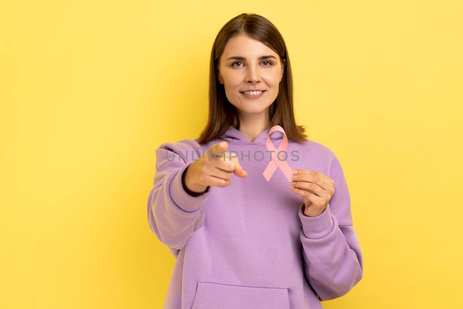 Smiling woman holding pink ribbon near her chest supporting another women, international symbol of breast cancer awareness, pointing to camera. Indoor studio shot isolated on yellow background