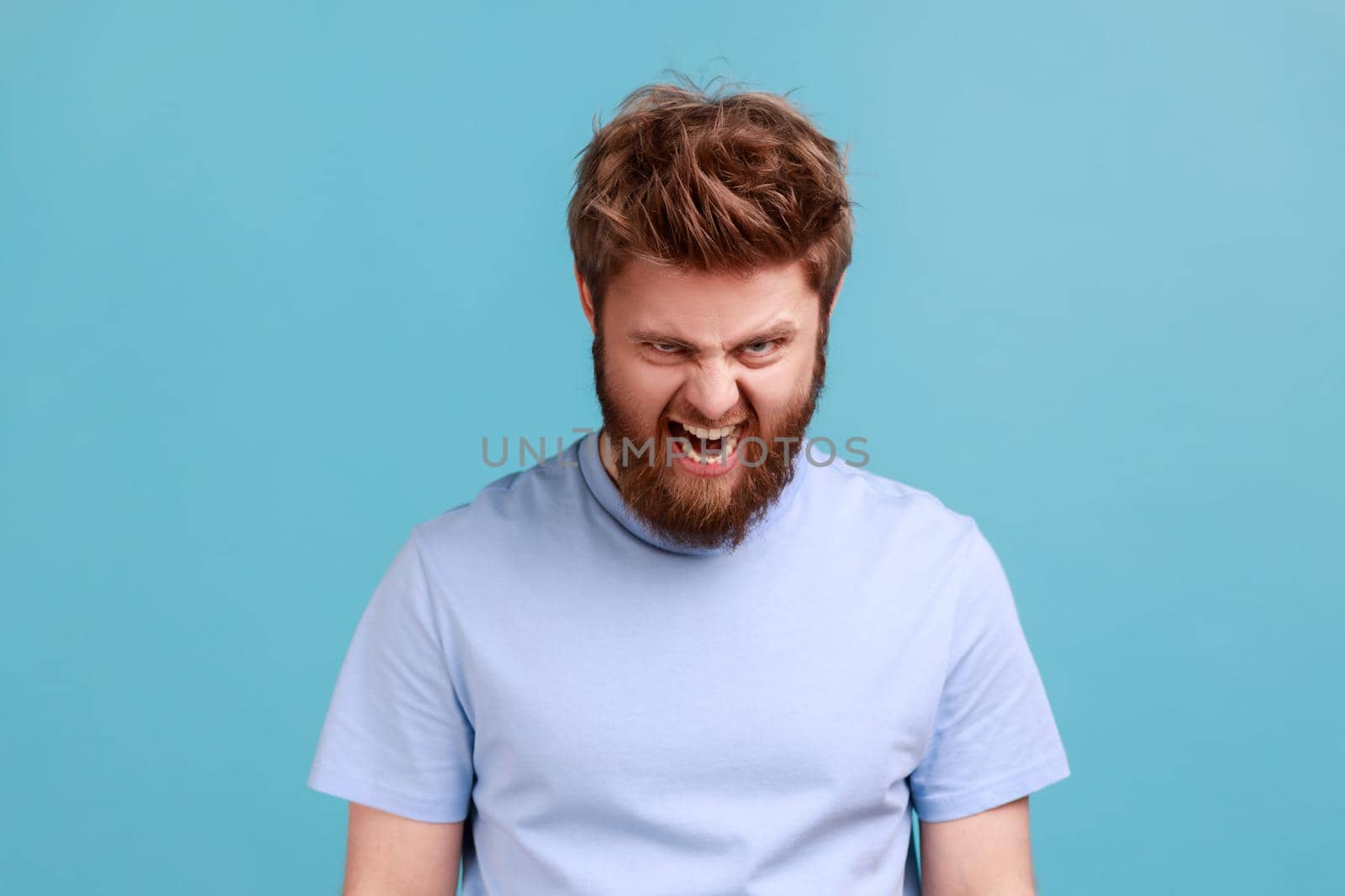 Portrait of bearded angry furious mad man standing and screaming with anger, expressing aggression, swearing with someone, having problems. Indoor studio shot isolated on blue background.