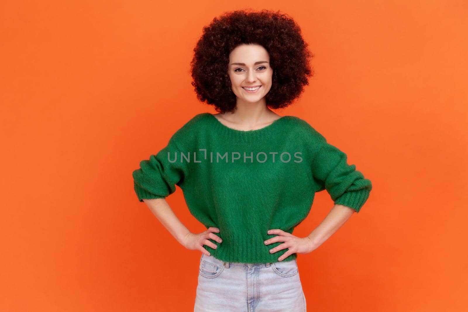 Good looking friendly confident woman with Afro hairstyle wearing green casual style sweater standing with hands on hips, smiling happily. Indoor studio shot isolated on orange background.