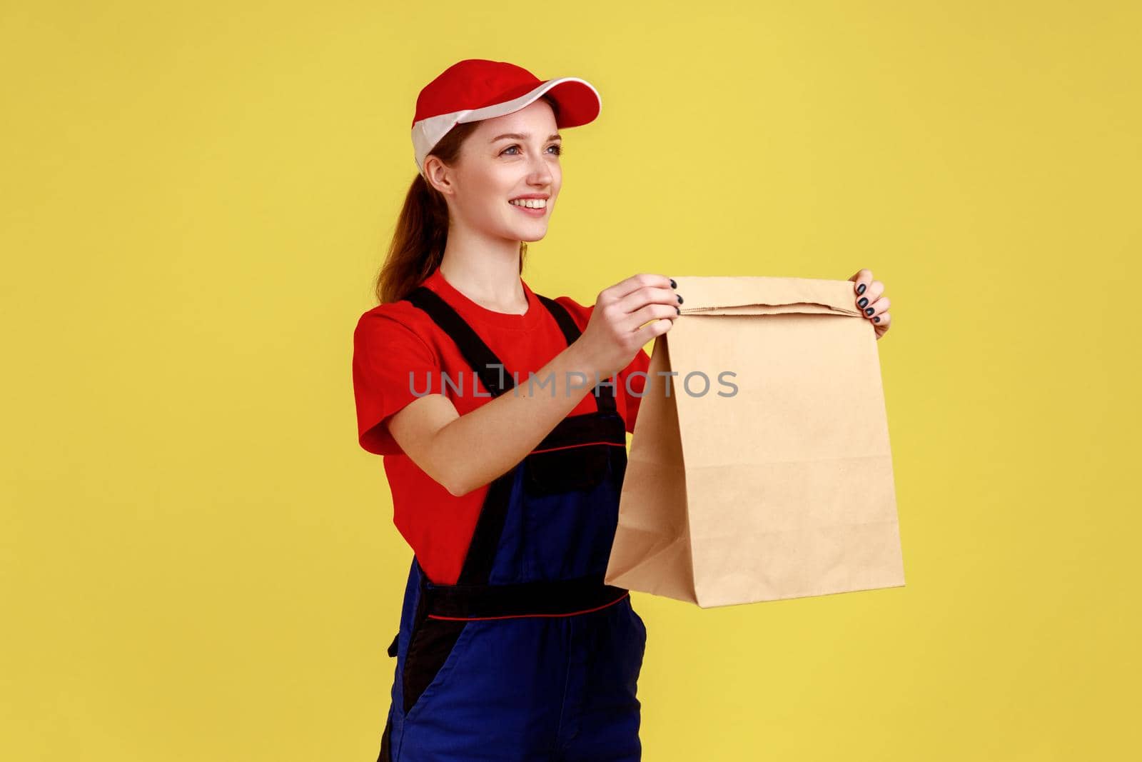 Portrait of positive delivery woman giving paper parcel with fast food, looking smiling at client, fast delivery, wearing overalls and red cap. Indoor studio shot isolated on yellow background.