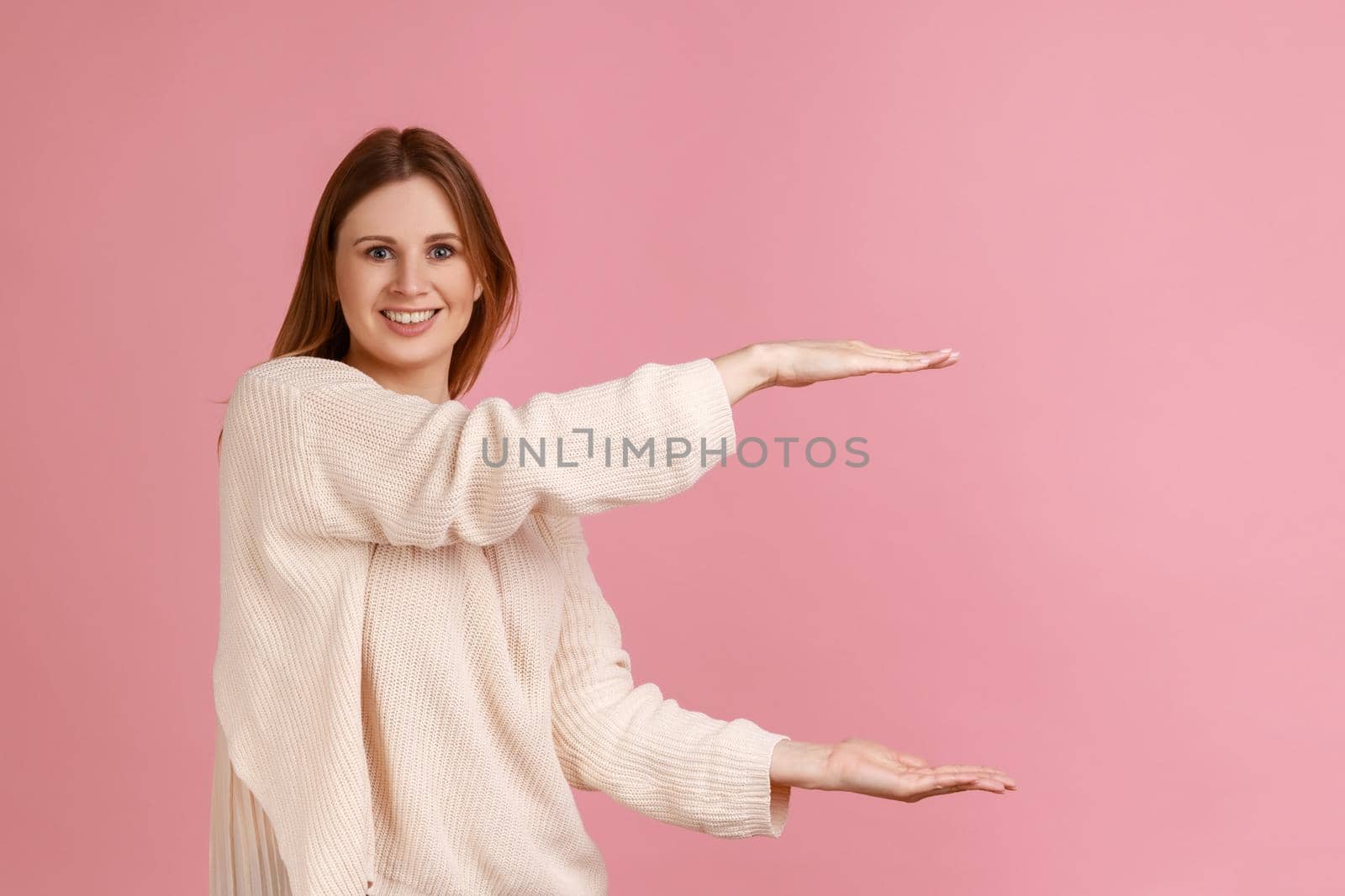 Portrait of happy smiling beautiful blond woman presenting area between hands for advertisement, showing huge size, wearing white sweater. Indoor studio shot isolated on pink background.