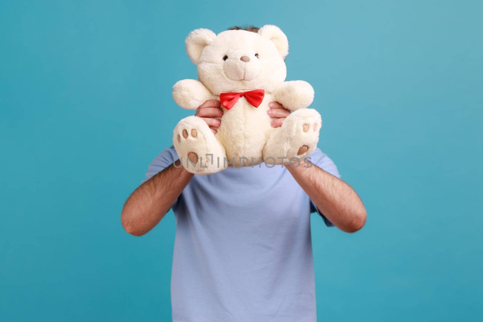 Portrait of unknown anonymous bearded man hiding his face behind white soft teddy bear, romantic surprise for his girflriend. Indoor studio shot isolated on blue background.