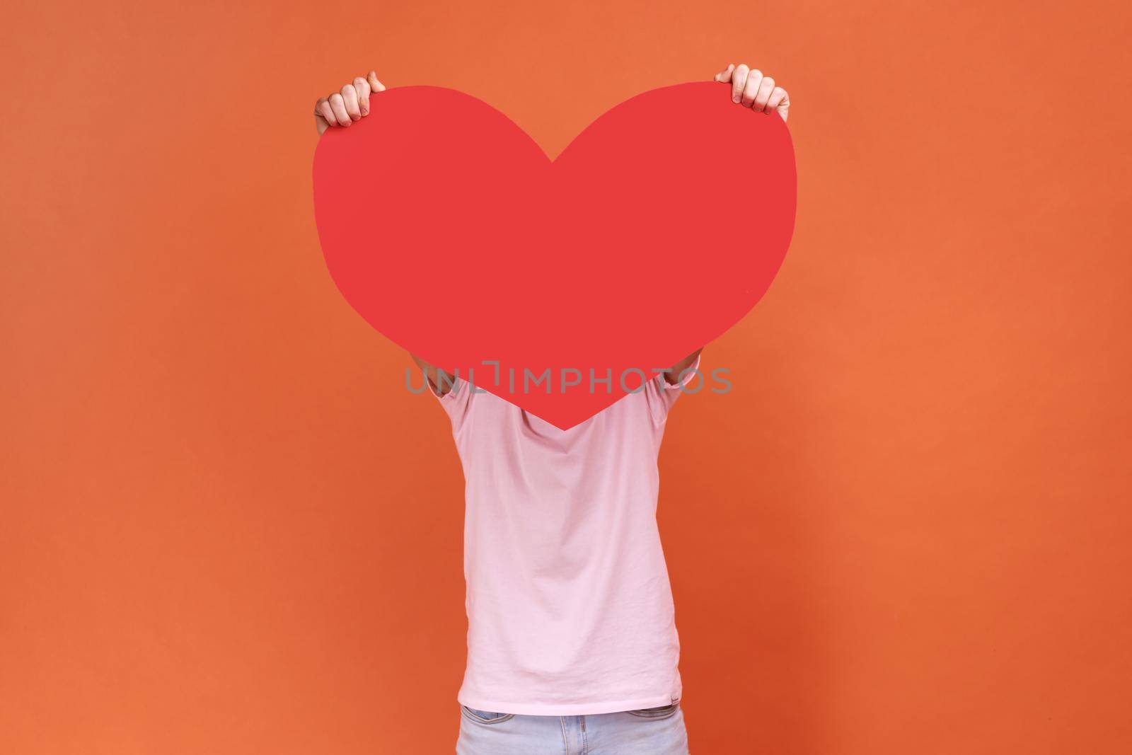 Portrait of anonymous bearded man hiding behind big red heart, holding symbol of love and affection, large greeting card, wearing pink T-shirt. Indoor studio shot isolated on orange background.