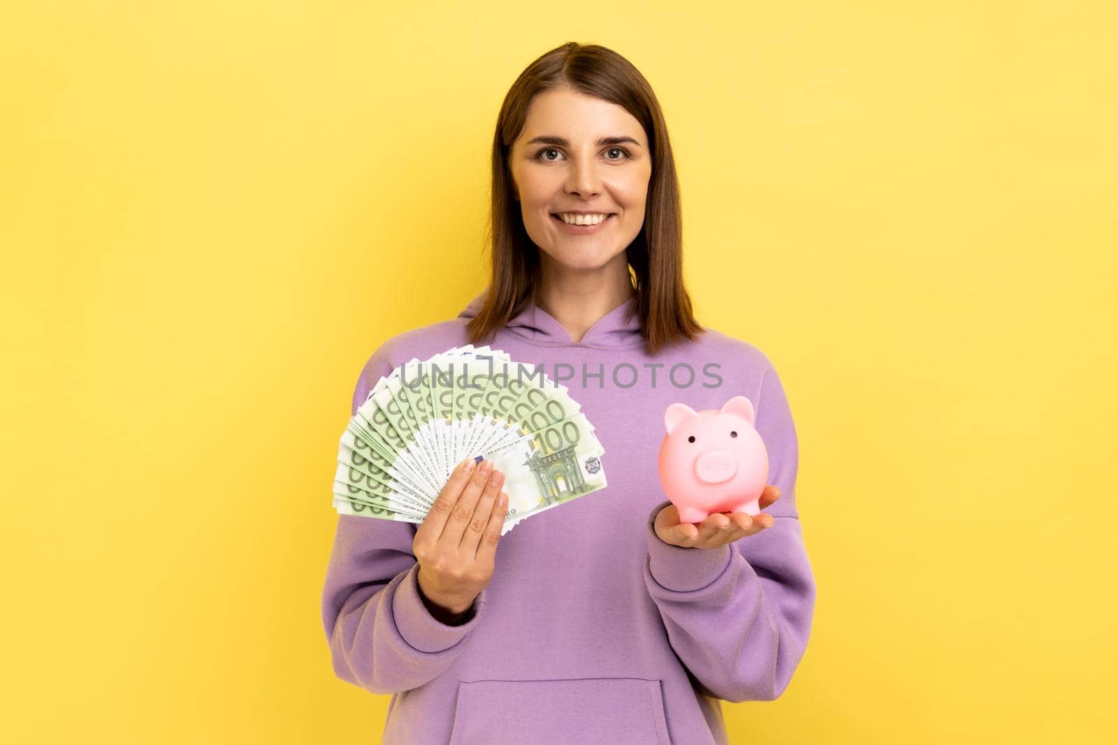 Smiling affirmative woman with dark hair holding big sum of money and piggy bank, profitable investment, wearing purple hoodie. Indoor studio shot isolated on yellow background.