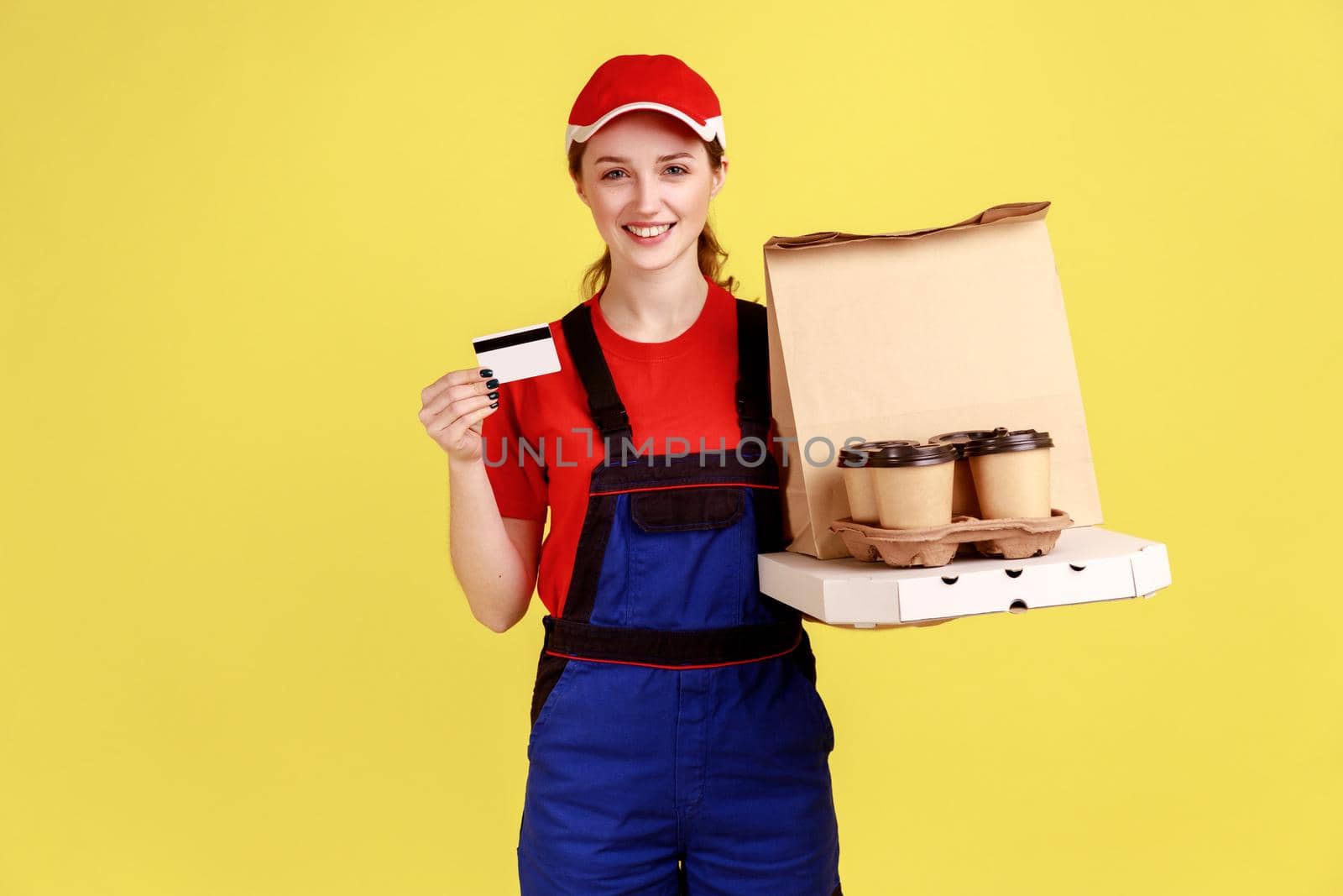 Portrait of optimistic courier woman holding credit card and coffee with pizza box, looking at camera with happy expression, wearing overalls and cap. Indoor studio shot isolated on yellow background.