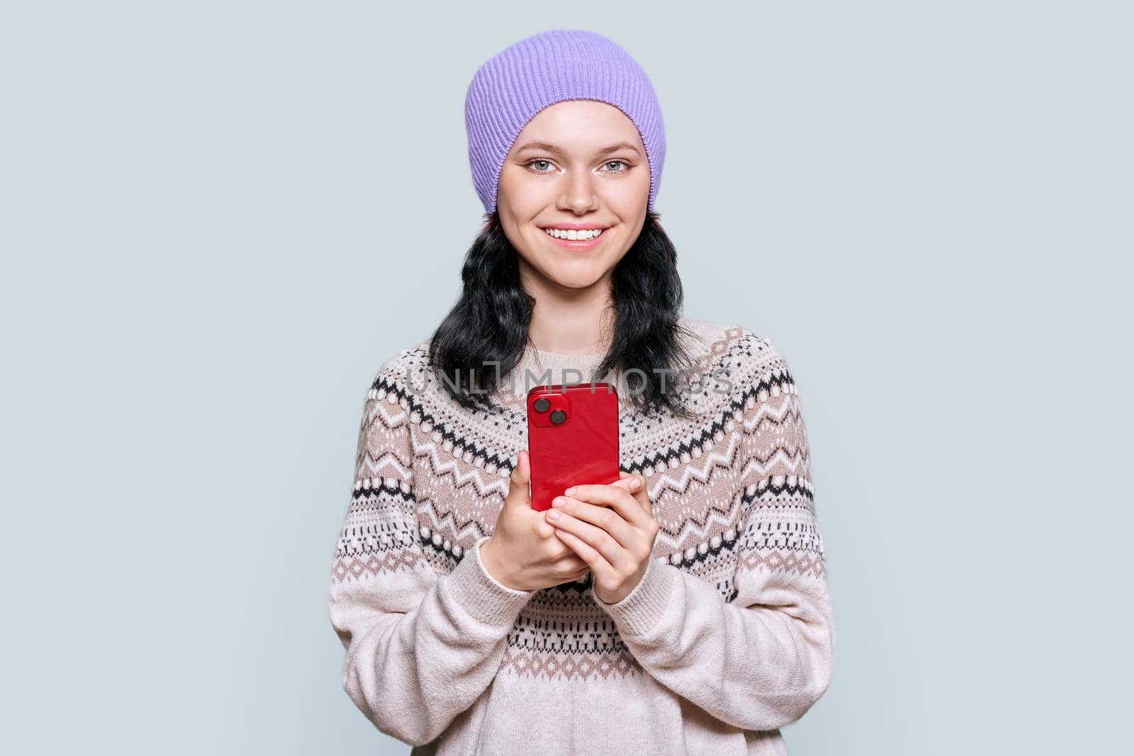 Beautiful young teenage girl in hat in a woolen sweater with smartphone in her hands looking at camera on light gray color studio background. Winter season, Christmas and New Year holidays concept