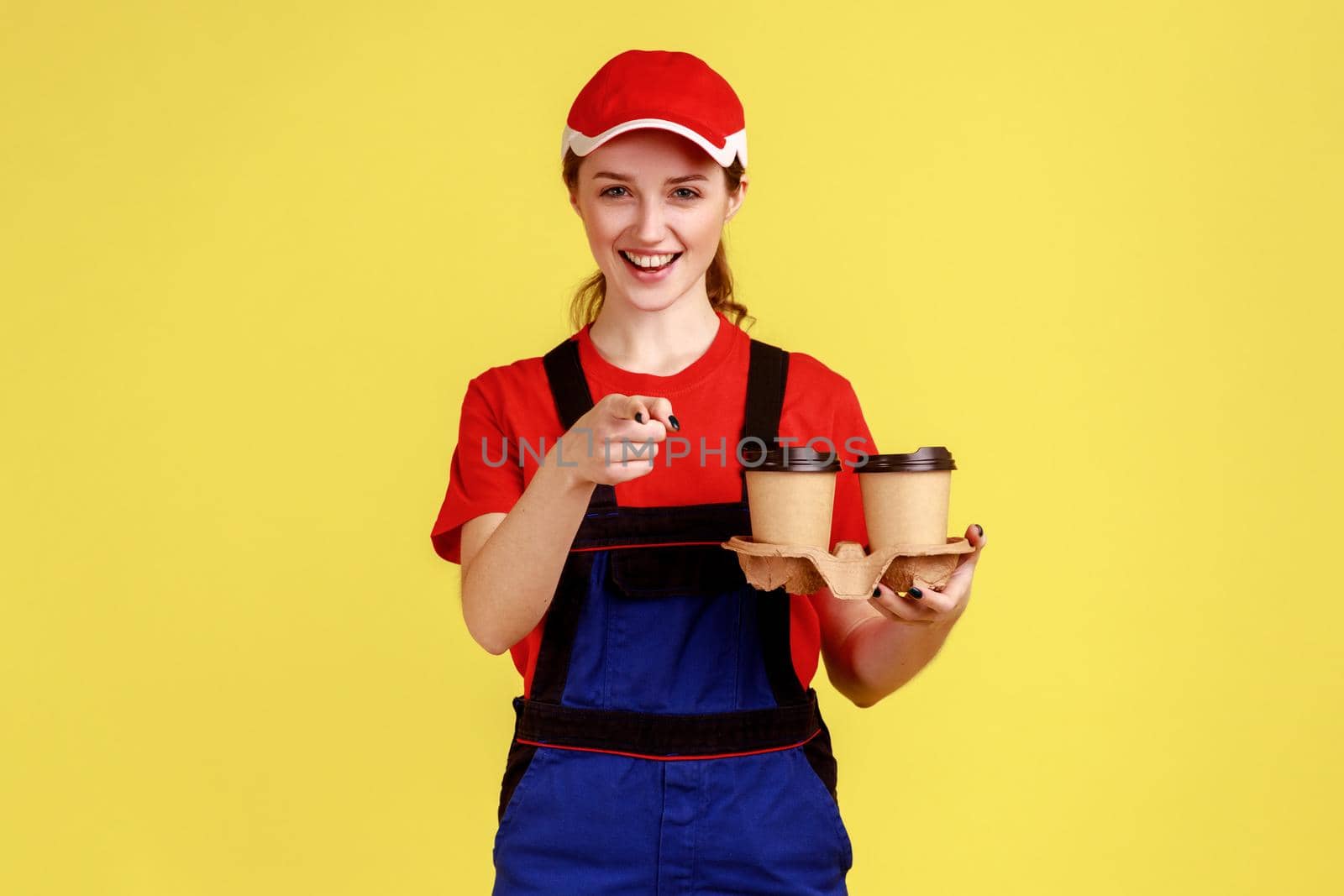 Portrait of delighted courier woman standing and holding coffee cups, pointing at camera, delivering beverage for you, wearing overalls and red cap. Indoor studio shot isolated on yellow background.
