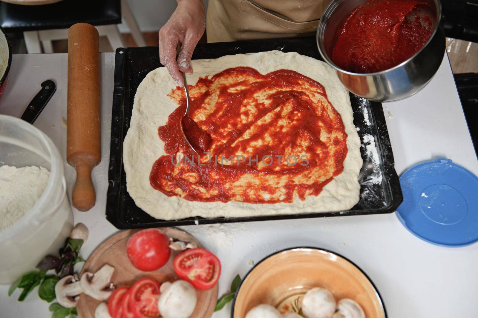 View from above. Housewife in beige chef's apron spreading freshly made tomato sauce on rolled out dough in pizza pan. Fresh organic ingredients on a kitchen table. Cooking. Culinary. Italian cuisine