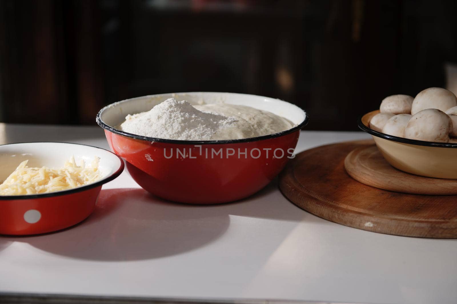 Enamel bowls with yeast rising dough, sprinkled with white flour, grated cheese and fresh mushroom champignons on a wooden cutting board on a white table in a country cottage kitchen. Food still life.