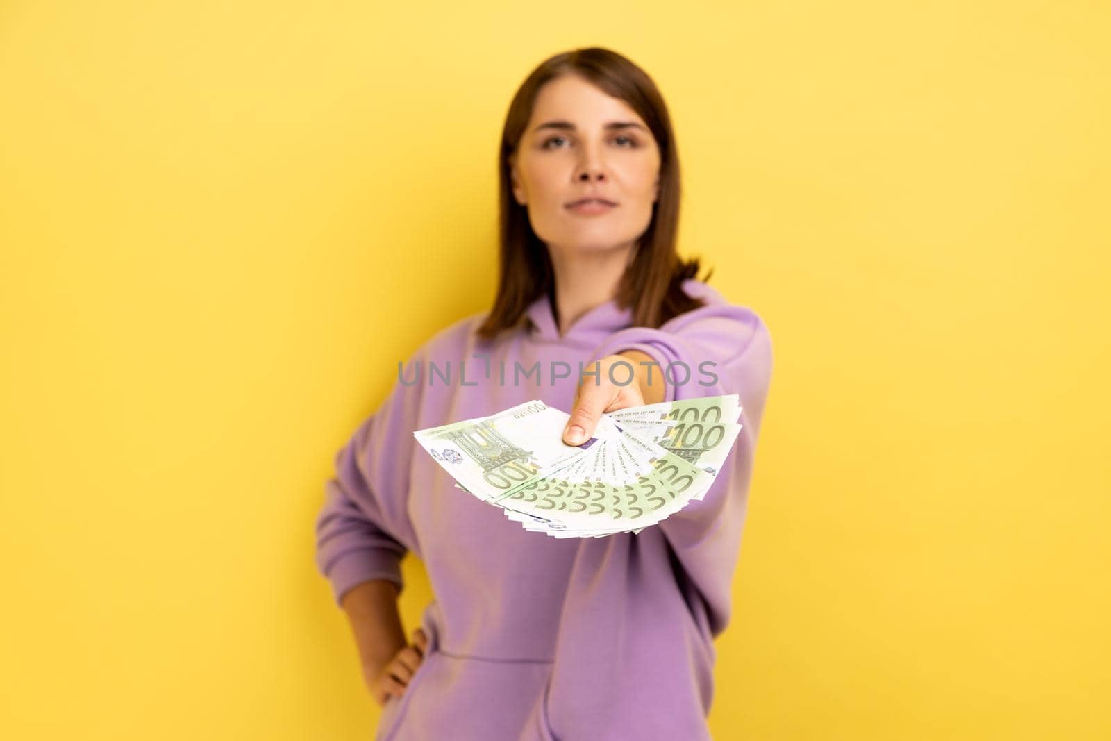 Portrait of rich confident young woman with dark hair holding out euro banknotes to camera, offering money, wearing purple hoodie. Indoor studio shot isolated on yellow background.