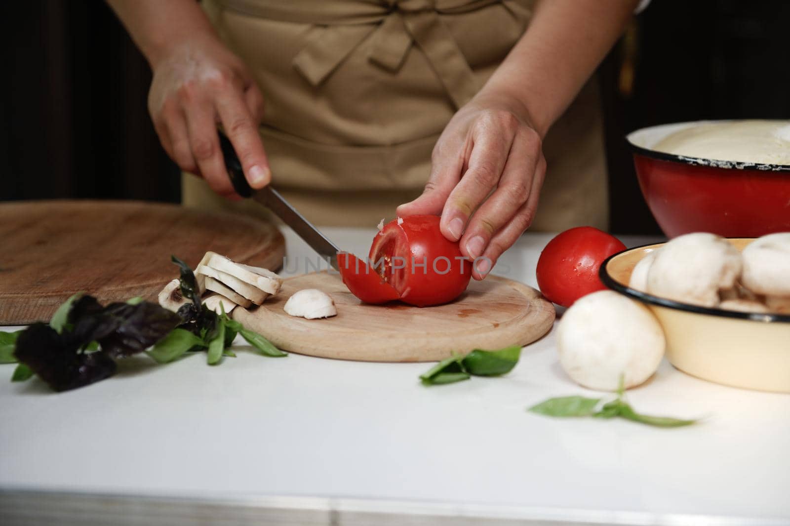 Close-up. Hands of a chef in a beige apron cut ripe juicy organic tomatoes on a chopping board with stacked sliced champignons and culinary aromatic herbs on the kitchen table, preparing healthy food