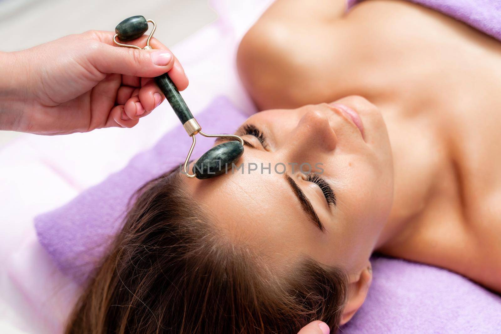 Relaxing massage. Side view european woman getting massage with jade face roller gouache in spa salon.