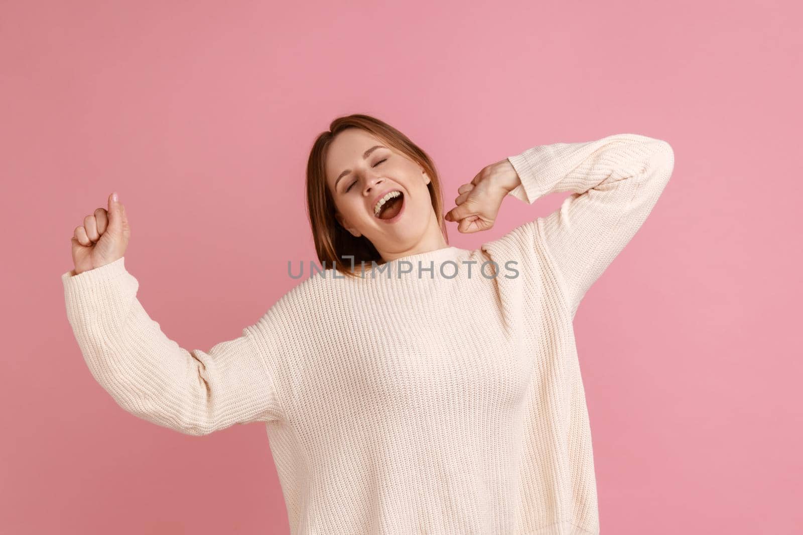 Portrait of sleepy young adult blond woman standing and yawning with closed eyes and raised arms, waking up early, wearing white sweater. Indoor studio shot isolated on pink background.
