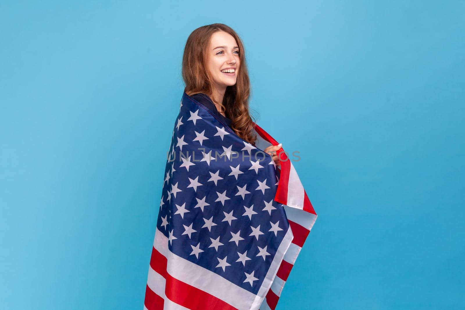 Side view of happy woman wearing striped casual style sweater, holding flag of united states of America, celebrating labor day, independence. Indoor studio shot isolated on blue background.
