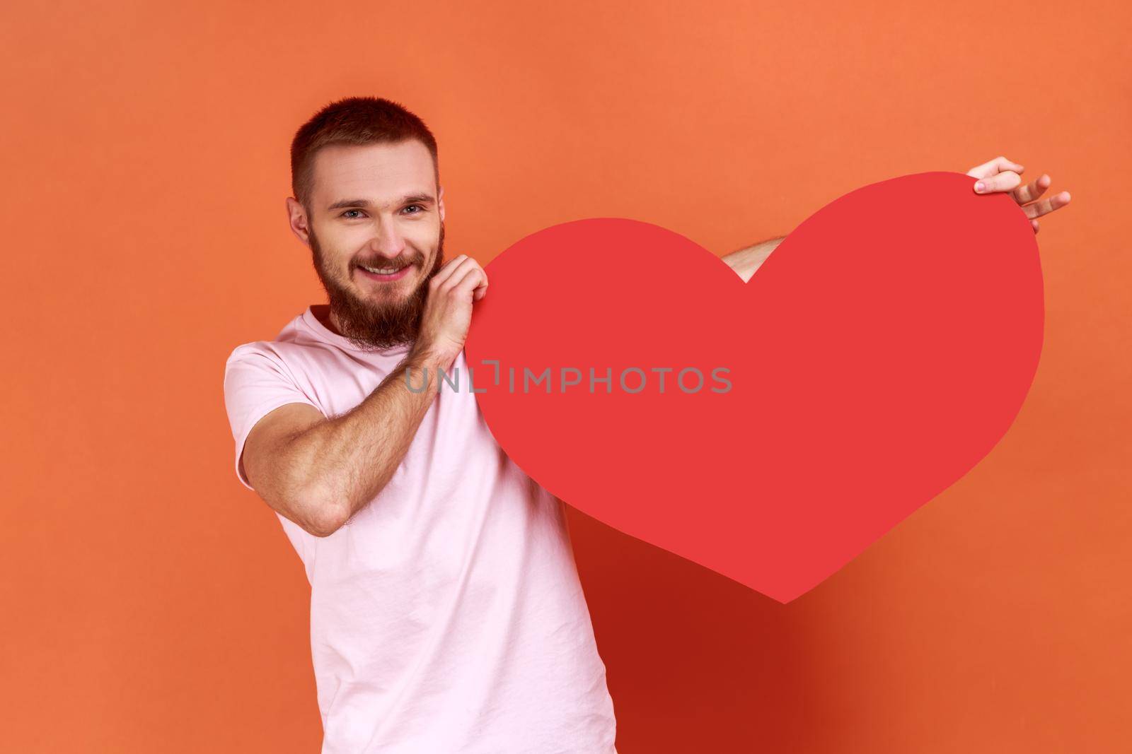 Portrait of smiling handsome bearded man holding big red heart, expressing positive romantic emotions, wearing pink T-shirt. Indoor studio shot isolated on orange background.