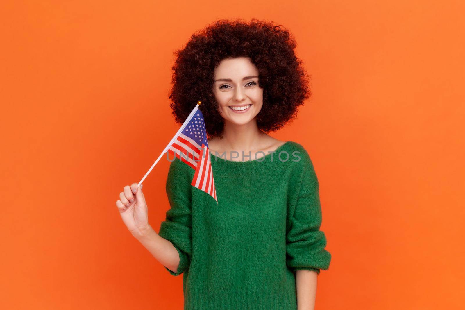 Happy positive woman with Afro hairstyle wearing green casual style sweater standing and waving american flag, celebrating national holiday. by Khosro1