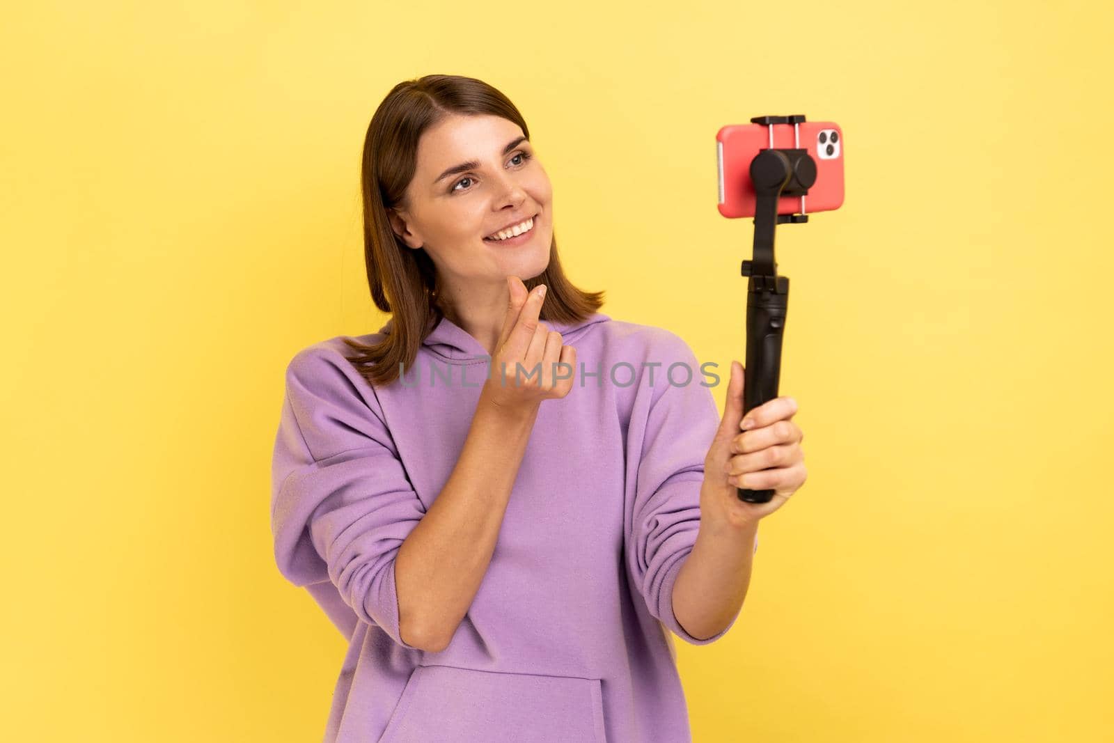 Optimistic woman using cell phone and steadicam for broadcasting livestream, showing korean finger heart sign, symbol of love, care, peace or support. Indoor studio shot isolated on yellow background.
