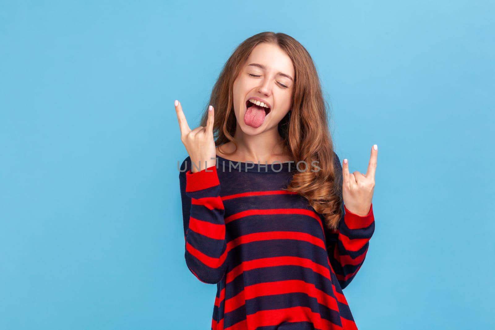 Yeah, awesome. Portrait of excited rocker woman in sweater showing rock and roll hand gesture, crazy devil horns with fingers and yelling. Indoor studio shot isolated on blue background.