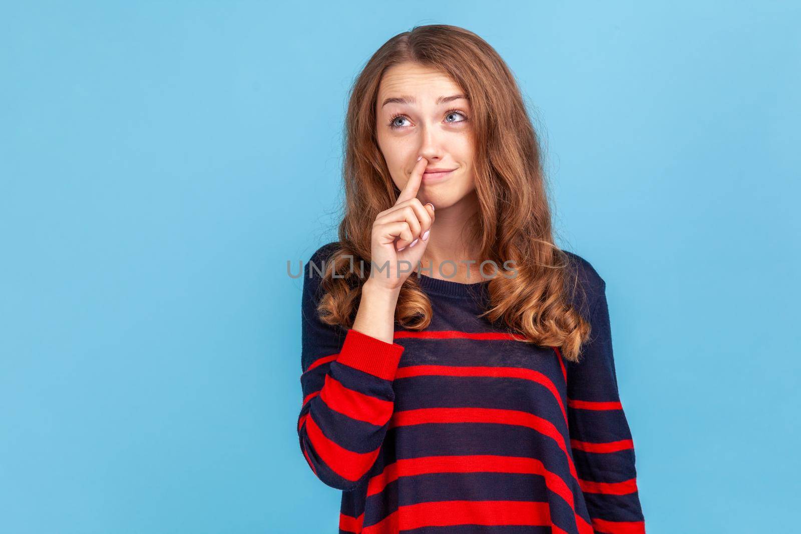 Childish woman wearing striped casual style sweater holding finger in her nose, uncultured bored girl having fun, bad manners, looking away. Indoor studio shot isolated on blue background.