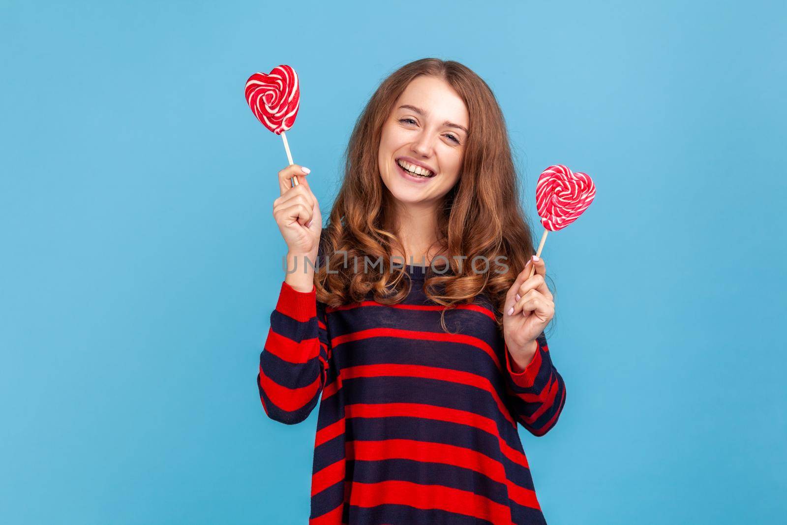 Satisfied young adult woman wearing striped casual style sweater, standing with two heart shape candies, looking at camera with toothy smile. Indoor studio shot isolated on blue background.