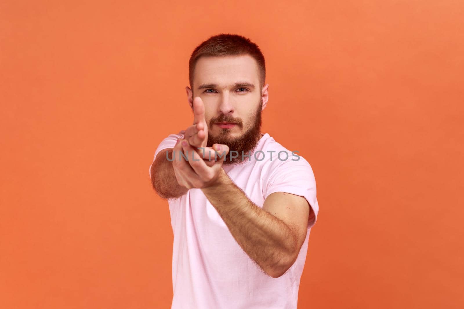 Portrait of serious brave bearded man holding fingers pretending holding gun, aiming enemy, self-defense, wearing pink T-shirt. Indoor studio shot isolated on orange background.