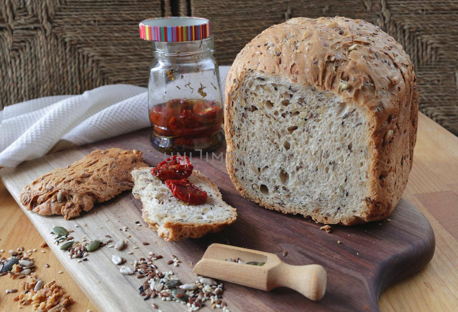 loaf of homemade whole grain bread and a cut slice of bread on a wooden cutting board. A mixture of seeds and whole grains. A jar of sun-dried tomatoes on a crust of fresh bread. Healthy food concept by Proxima13