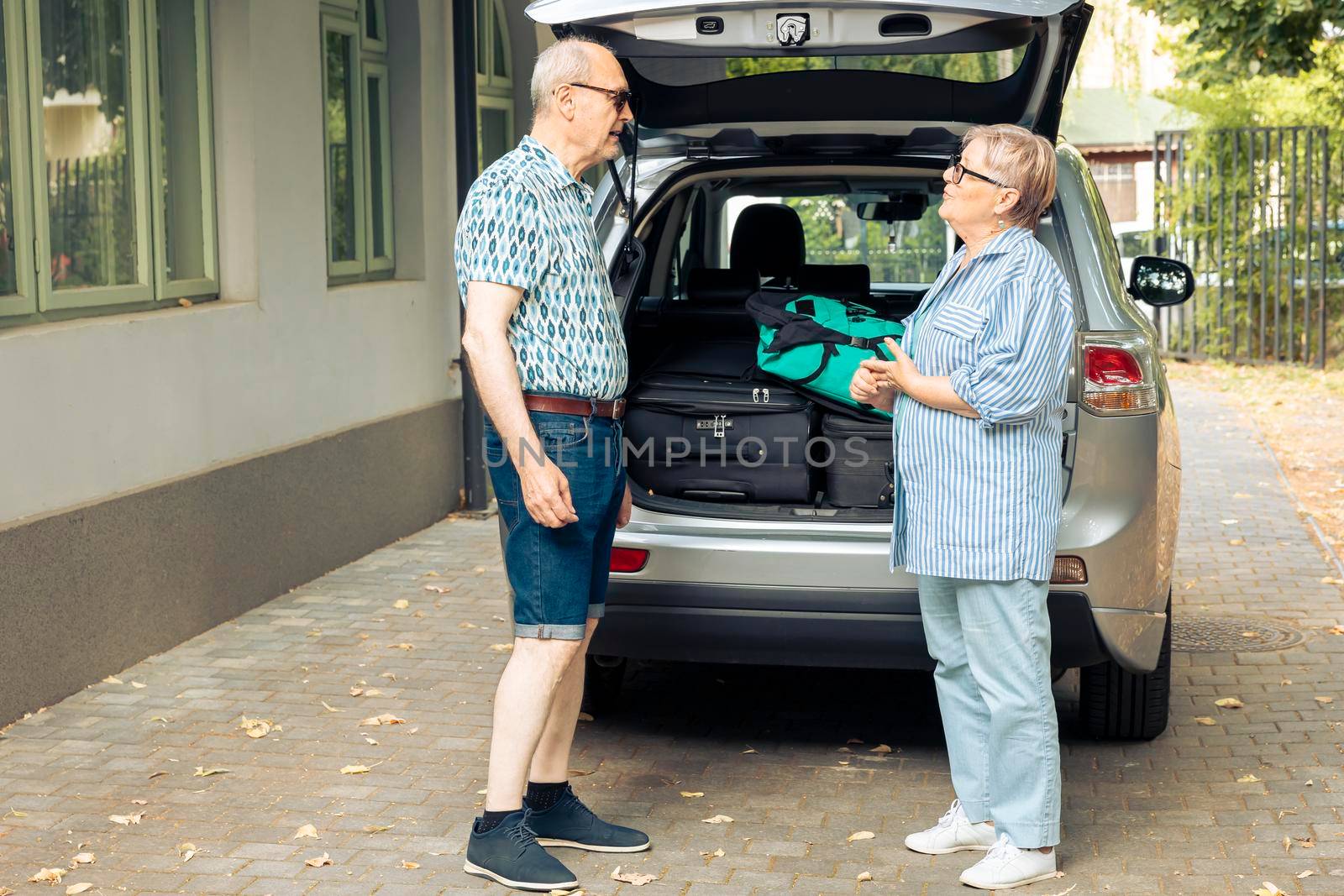 Aged people going on holiday vacation, loading suitcase and luggage in automobile trunk at home. Travelling by car with baggage and bags, leaving onubran cityscape adventure trip.