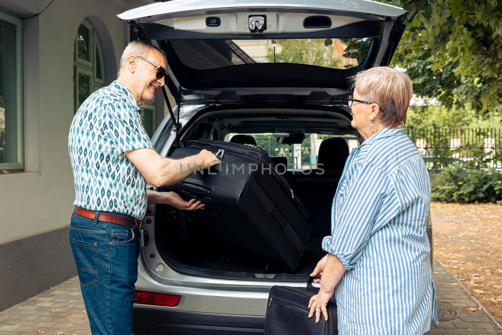 Aged couple putting bags in vehicle trunk to leave on retirement holiday trip during summer. Travelling on road trip for recreation, packing baggage and suitcase for urban cityscape vacation.