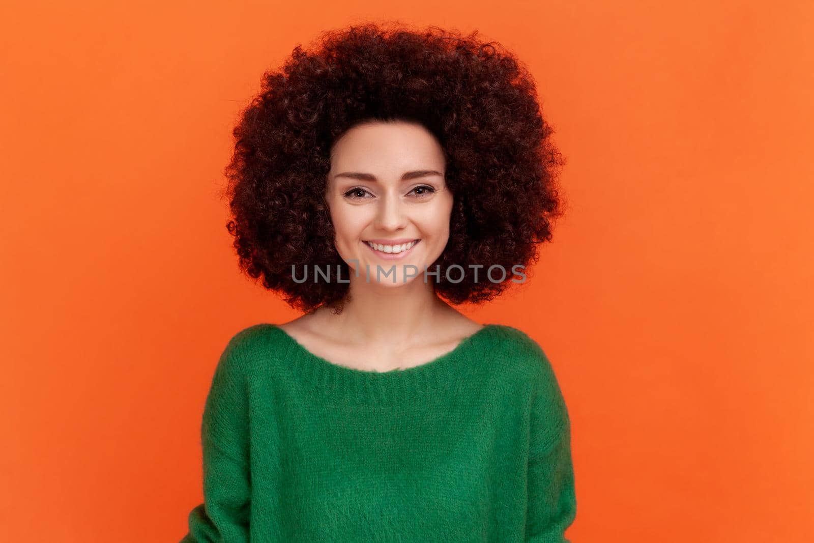 Portrait of beautiful positive woman with Afro hairstyle wearing green casual style sweater looking at camera, has pleasant emotions. Indoor studio shot isolated on orange background.
