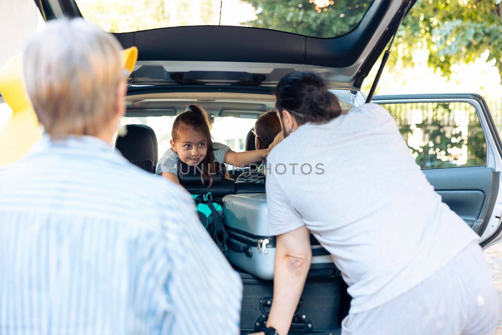 Young kid in vehicle leaving on vacation with parents and grandparents, loading travel bags in trunk of automobile. Big happy family travelling on road trip holiday to go to seaside destination.