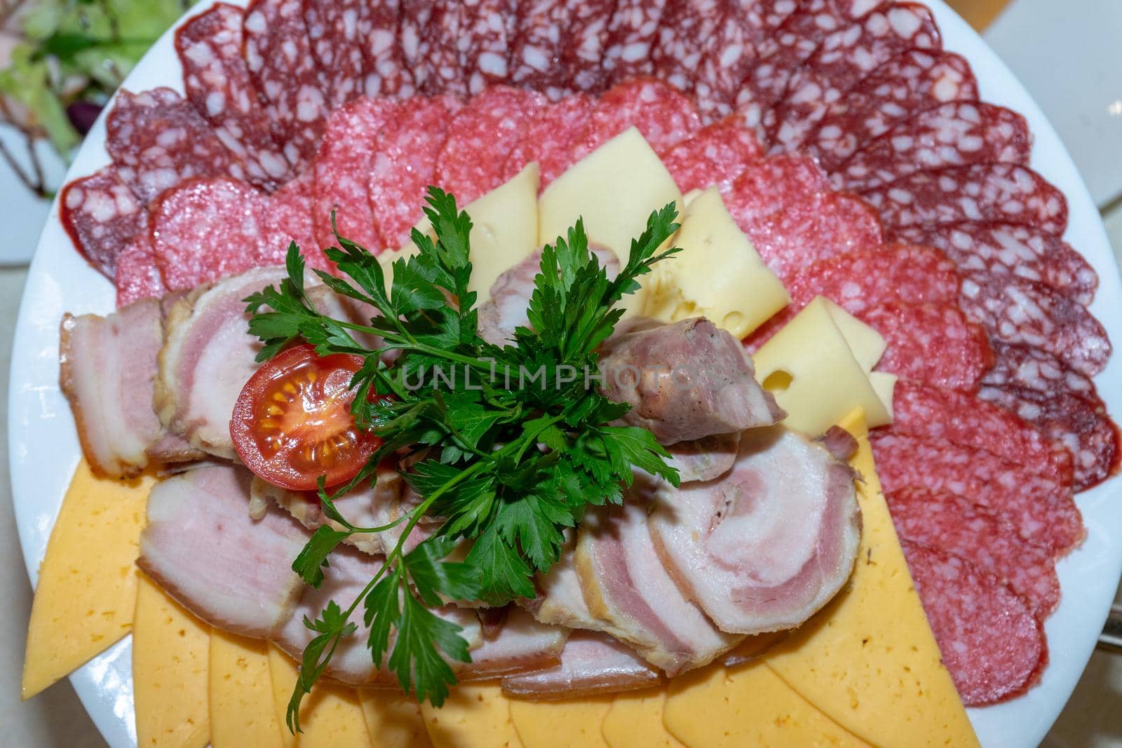 A plate of cold appetizers with various sausages, meat, and cheese. Greens and tomatoes