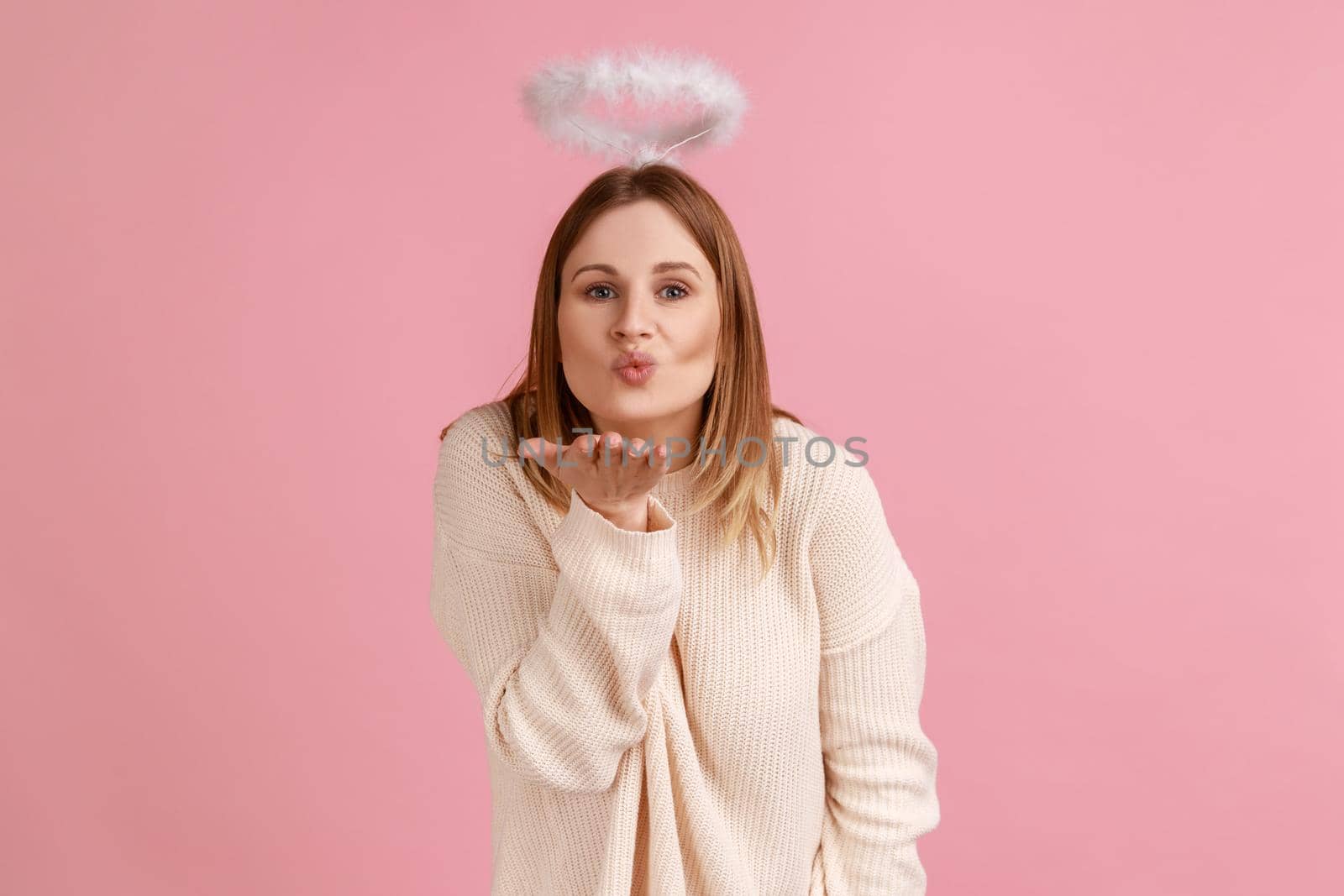 Portrait of romantic charming blond angelic woman with nimbus over head looking at camera and sending air kisses, wearing white sweater. Indoor studio shot isolated on pink background.