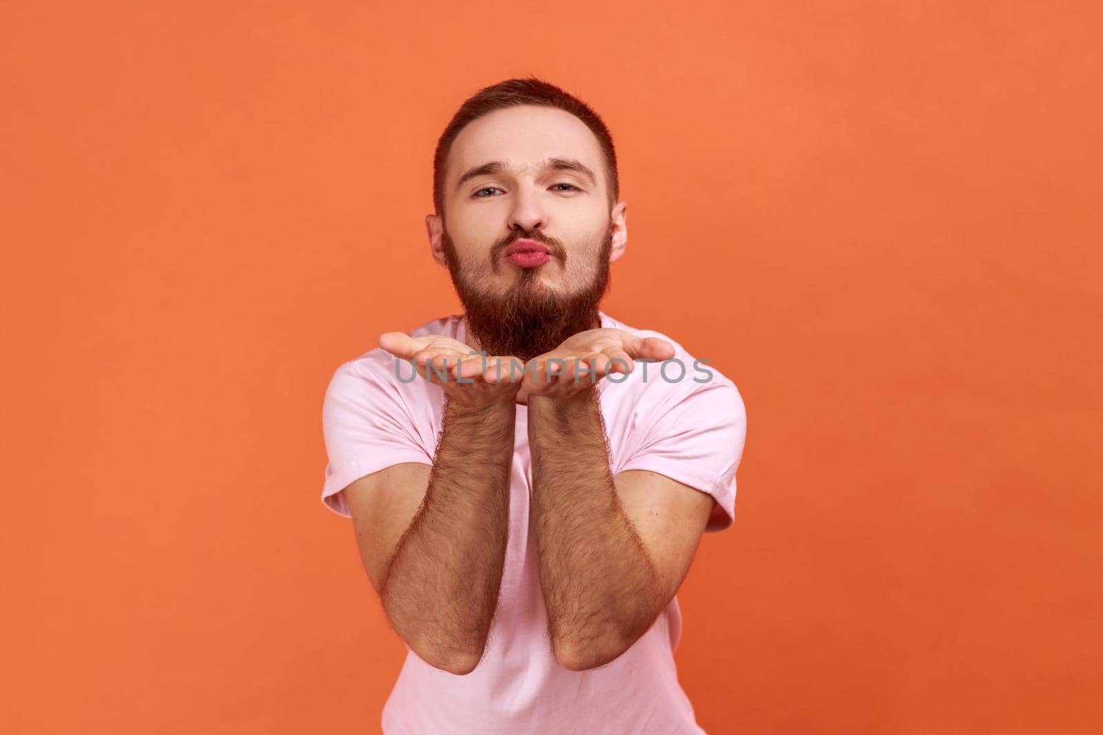 Portrait of handsome romantic young adult bearded man standing and looking at camera and sending air kiss, wearing pink T-shirt. Indoor studio shot isolated on orange background.