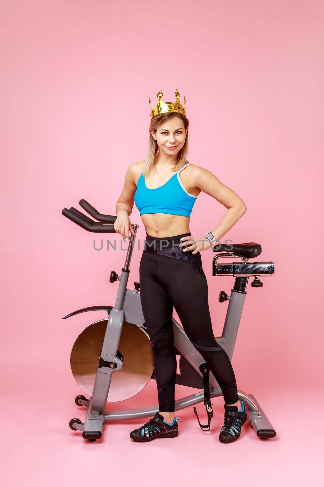 Full length of athletic woman in golden crown, queen of sports trainings, standing with hand on hips near bike simulator, wearing sports tights and top. Indoor studio shot isolated on pink background.