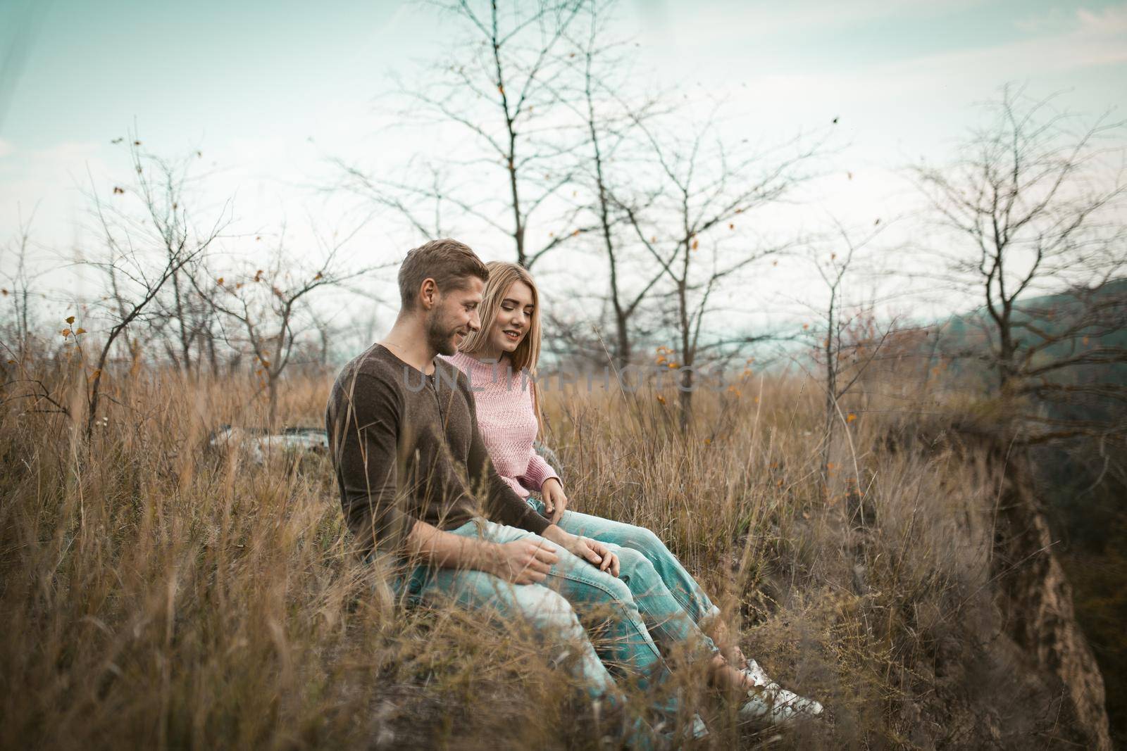 Happy Man And A Woman Are Sitting On The Edge Of A Cliff With Their Legs Dangling Down. Couple Of Tourists Sitting On The Grass On A Hillside In The Fall In The Fresh Air Outdoors