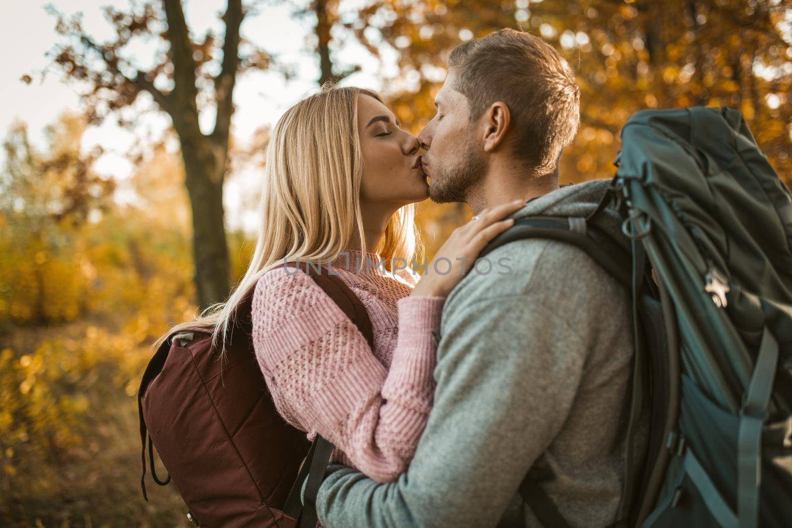 Couple Of Tourists Kissing in Forest Outdoors, Man And Woman Hug And Kiss Standing Outdoors With Yellow Trees On Backgroung, Portrait Of Cheerful Backpackers Standind In In Sunlight