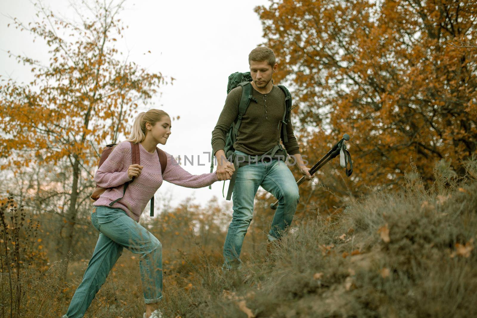 Loving couple of tourists climb the slope holding hands. Cheerful Young Couple, man and a woman with backpacks travel together in autumn nature. Hiking concept. Healthy lifestyle concept.