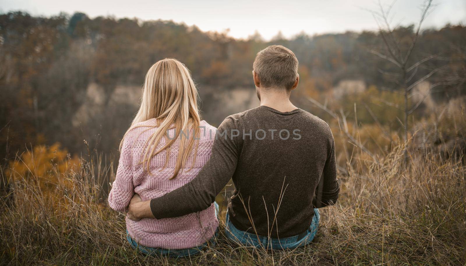 Couple Sits Embracing On Cliff Edge, Rear View Of Couple In Love Admiring Sunset Outdoors, With Autumn Forest And Cloudy Sky On Blurred Background