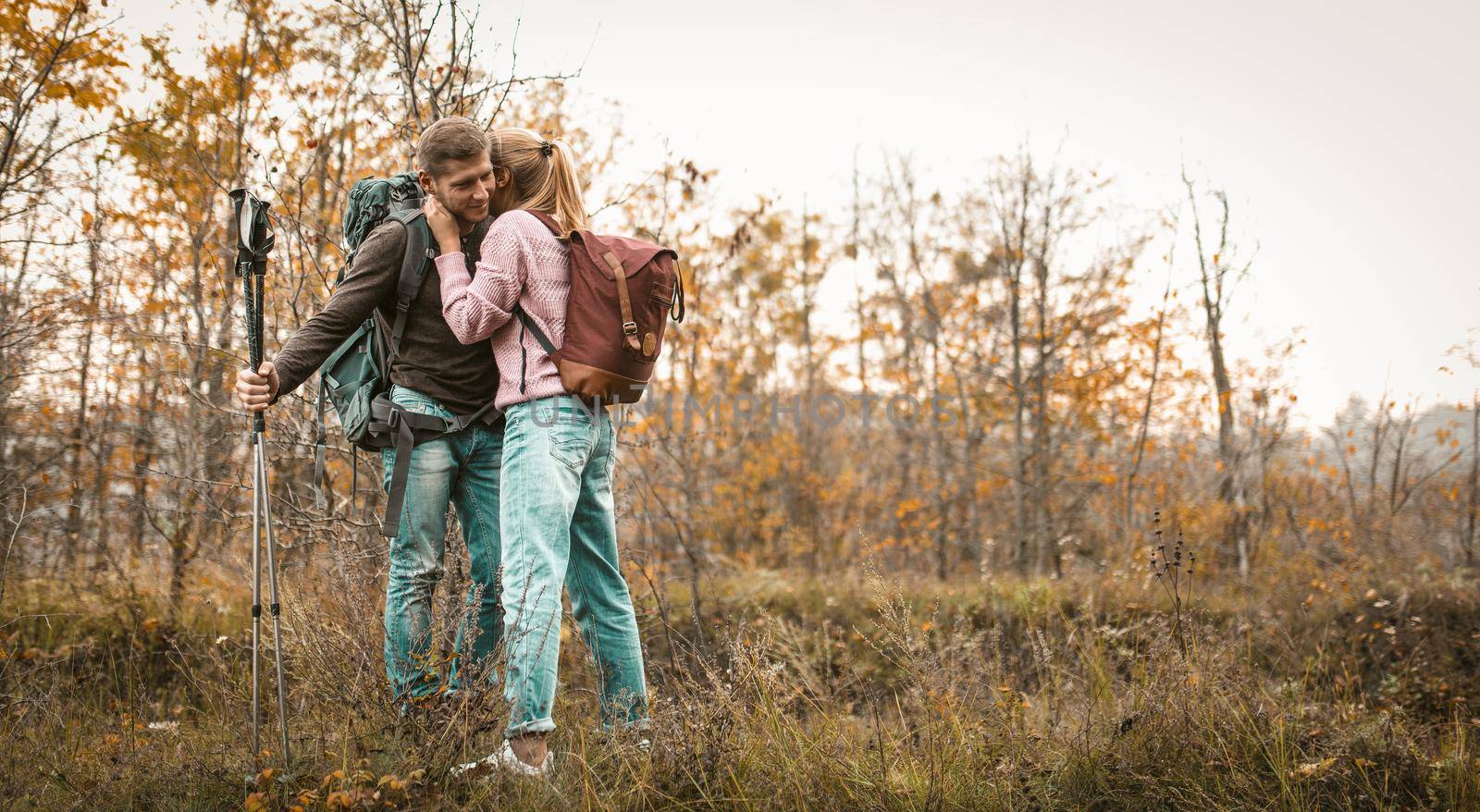 Traveling Couple Stopped To Relax In A Forest Glade, A Blonde Hugged Her Husband Kissing Him Or Saying Something In His Ear, Man Stands Leaning On Hiking Pools