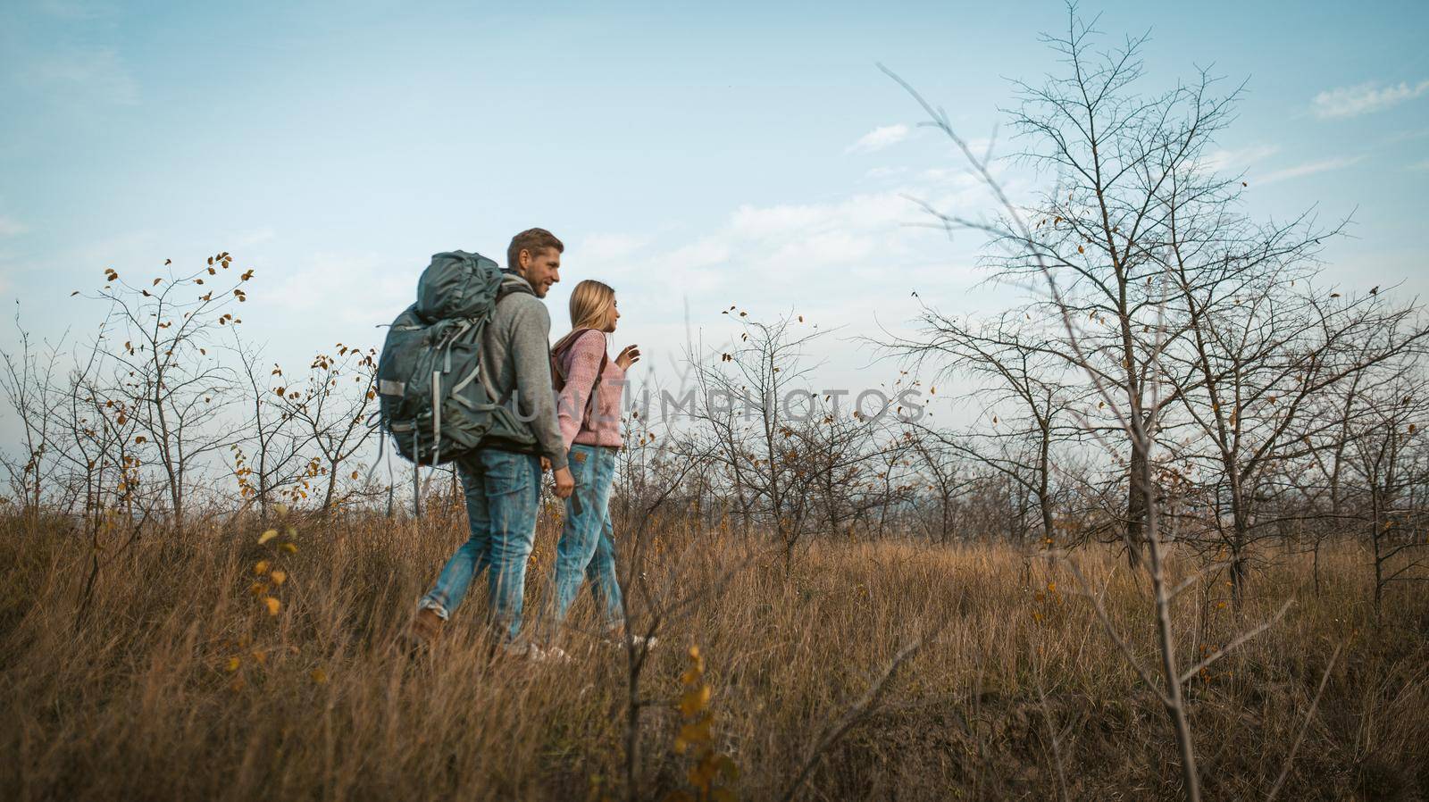 Couple of travelers walking through an autumn field holding hands. Tourists at full height against a background of blue sky and autumn natural landscape. Shot from below. Hiking concept by LipikStockMedia