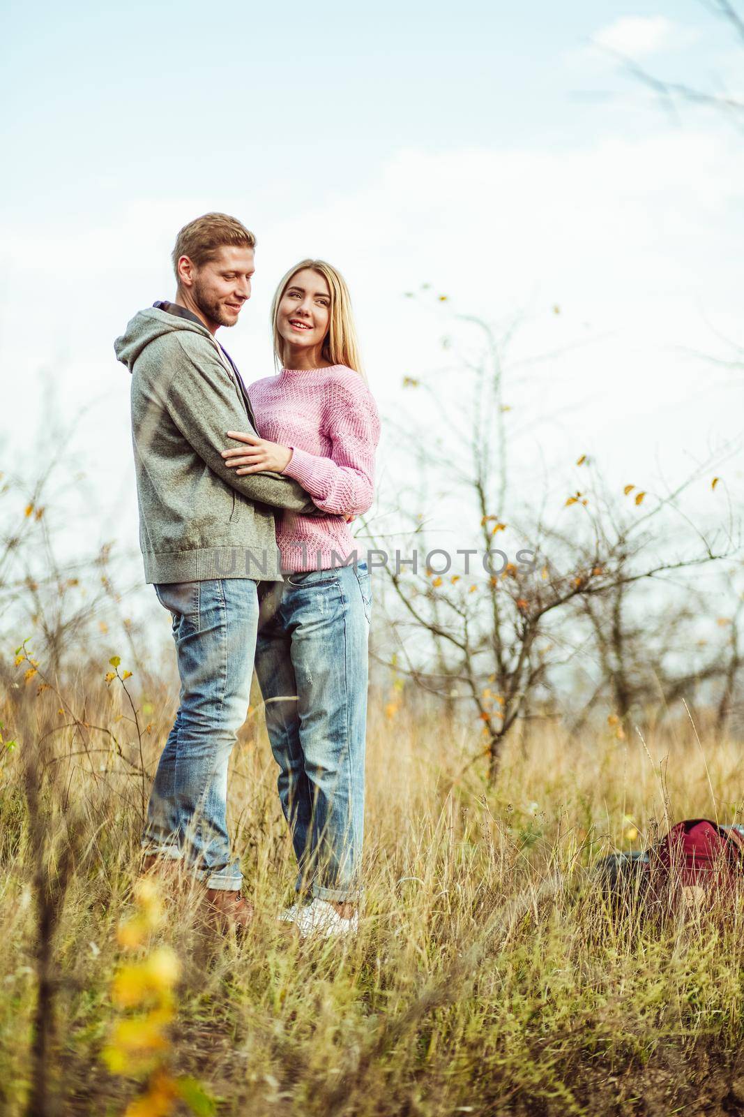 Hugs in nature. Young woman and man stands hugging in the field against evening sky. Couple of tourists in full growth.