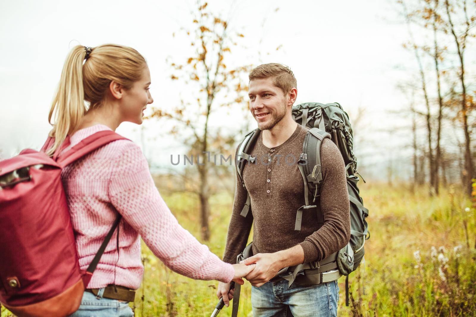 Romantic date for couple of backpackers in wild nature. Happy man and women looks each other holding hands while standing against background of autumn field. Hiking concept. Adventure concept by LipikStockMedia