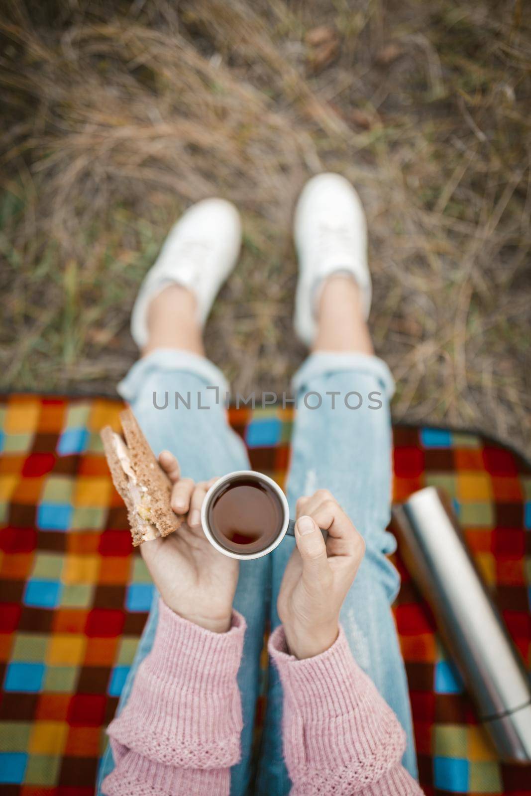 Picnic or snack in nature. Female hands holding a cup of tea and a sandwich. Young woman or teen girl snacking while sitting on grass. Close-up shot. High angle view.