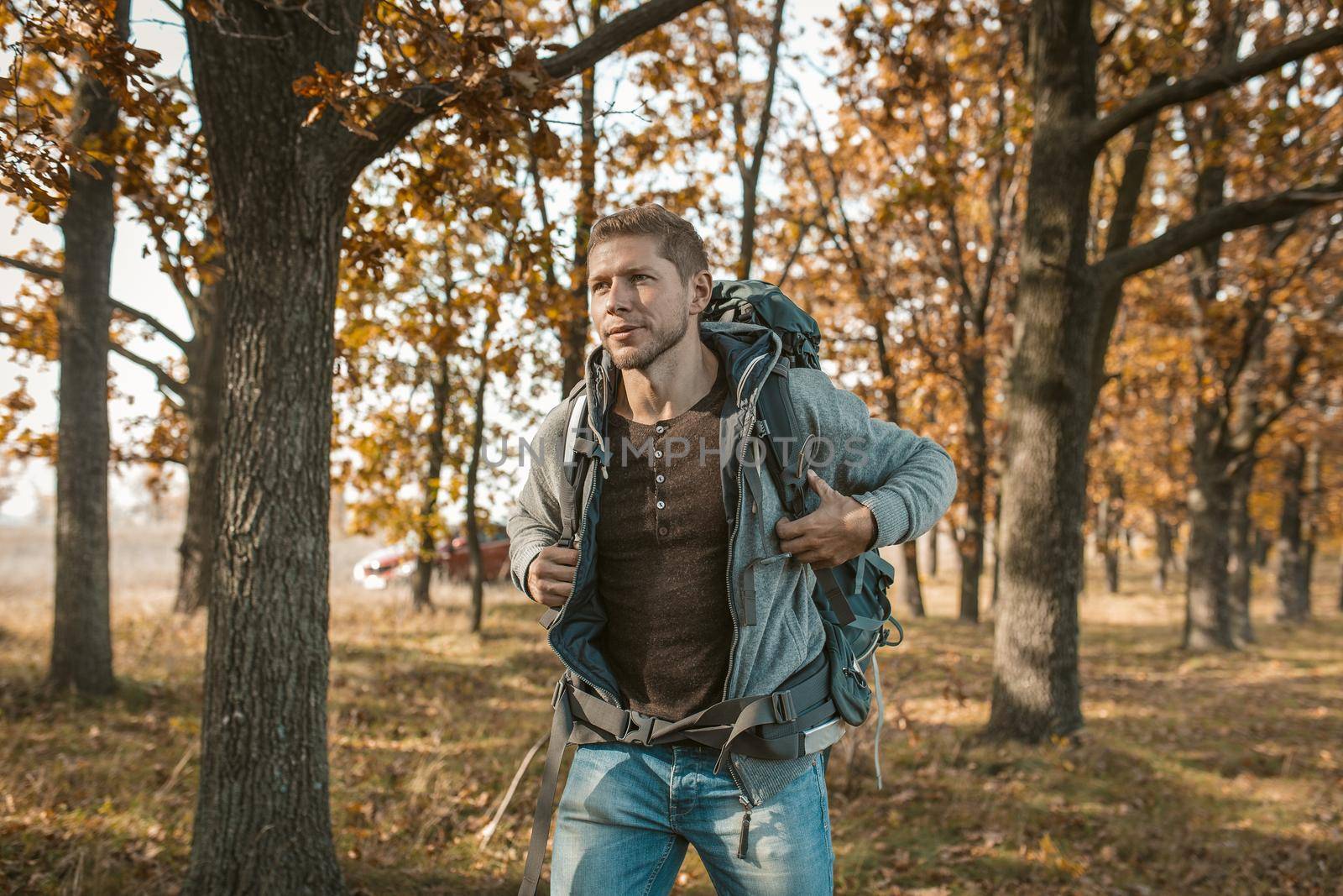 Tourist at the beginning of the journey. An inspired guy with a big backpack got out of a red car and begins his journey through the autumn forest. Hiking concept.