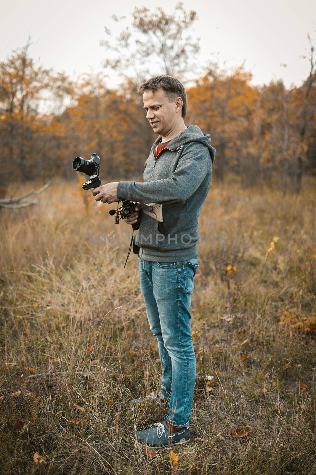 Positive camera man preparing camera on digital stabilizer for shooting photo and video content. Caucasian journalist or Paparazzi prepare for shooting while standing on grass among autumn nature.