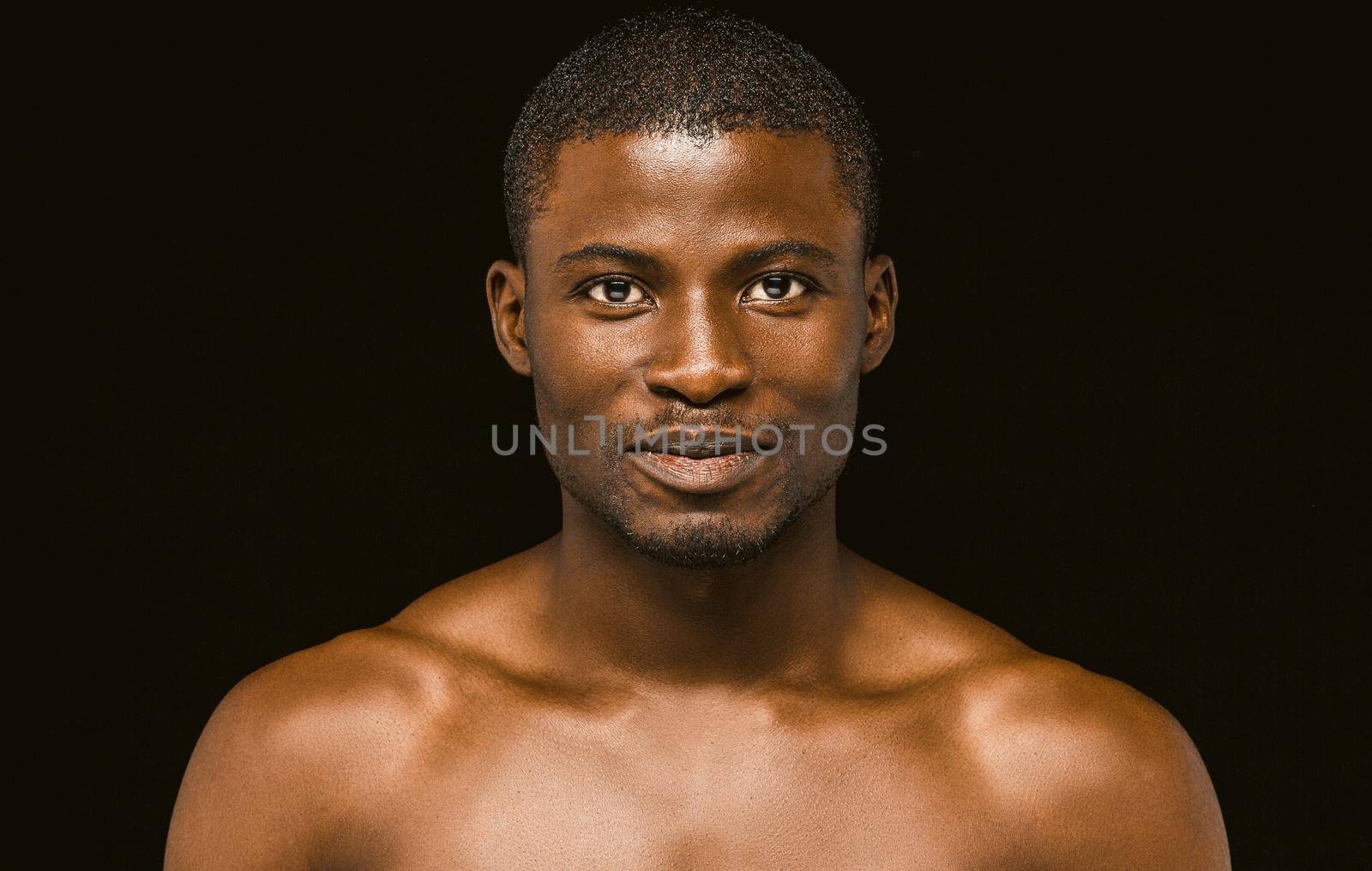 Nude smiling African macho man posing on black background, portrait of an attractive African american man smiling slightly while looking at the camera. Toned image by LipikStockMedia