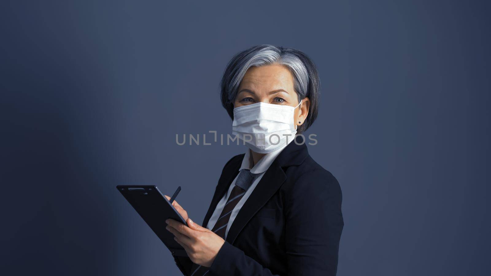 Gray haired businesswoman in protective mask smiles looking at camera on gray background. Pandemic concept.