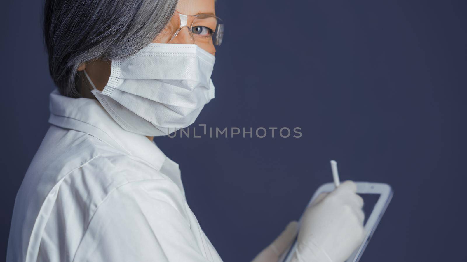 Woman in medical uniform holds digital tablet looking at camera. Grey haired female doctor on blue gray background with copy space at right side. Close up shot. Tinted image.